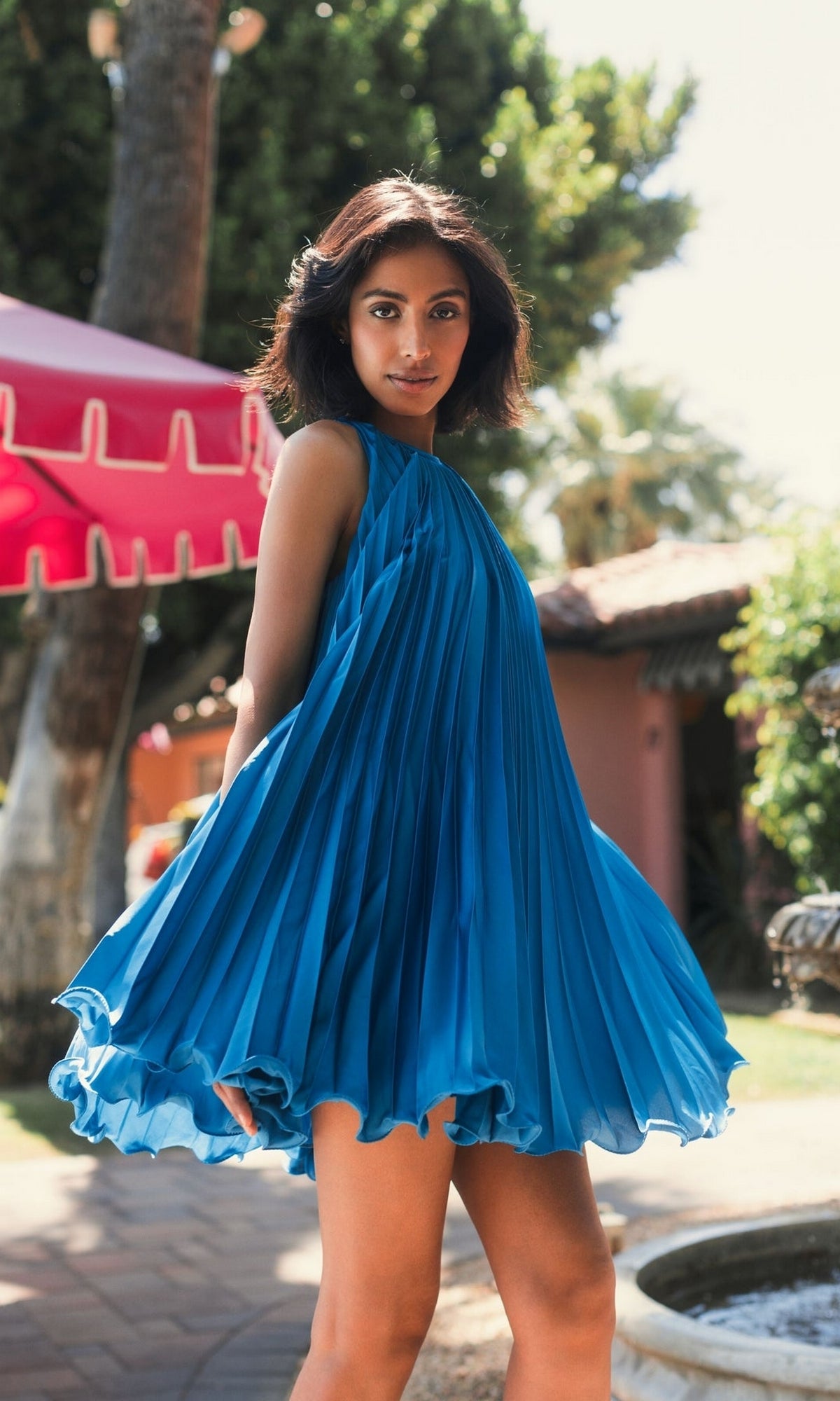 A woman stands outdoors in sunlight, wearing the Mac Duggal 49636 short pleated semi-formal dress in blue; greenery and a red canopy appear in the background.